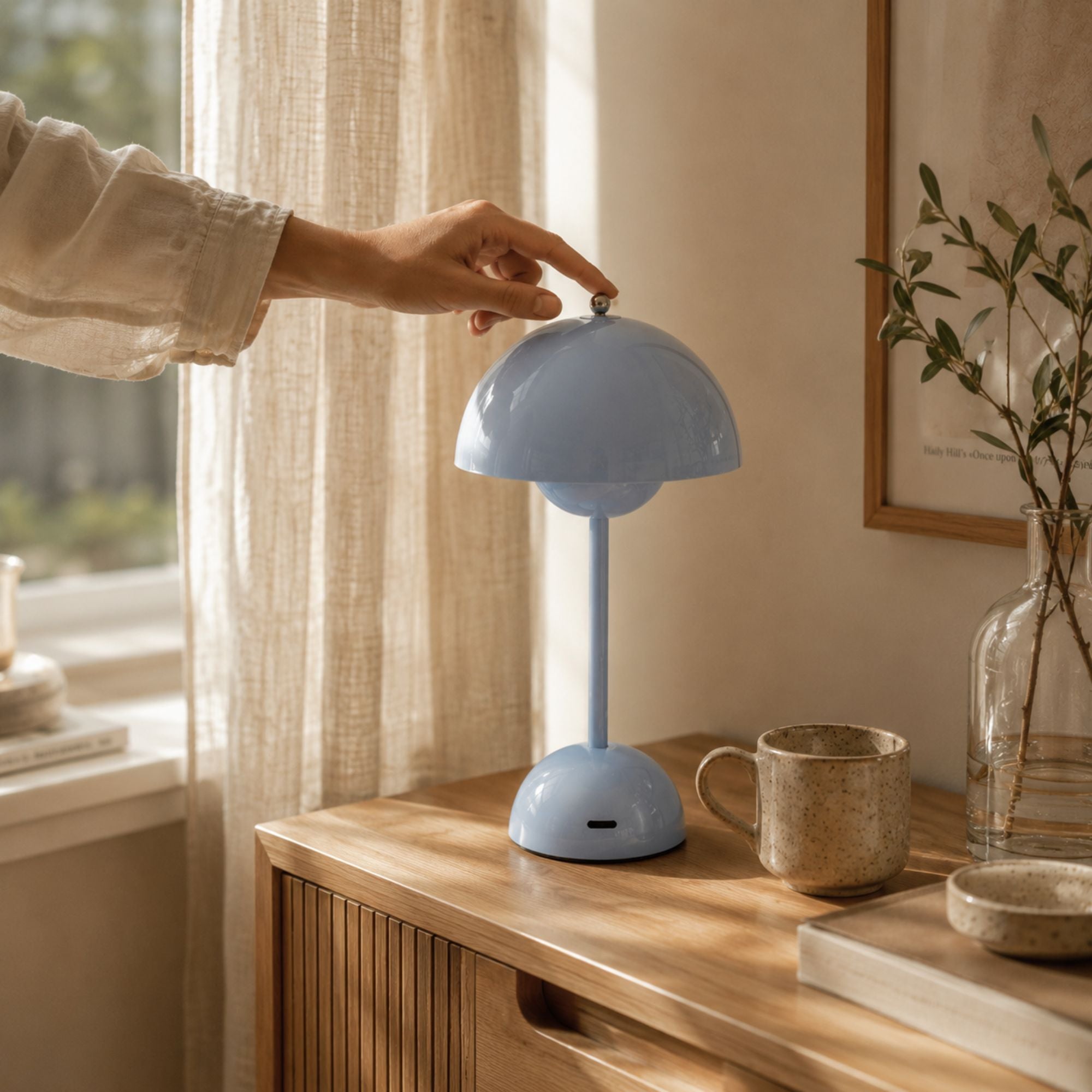 Ljusblå uppladdningsbar bordslampa med touchfunktion på ekmöbel i skandinavisk inredning. En hand tänder lampan intill keramikmugg, glasvas och linnégardiner i varmt dagsljus.