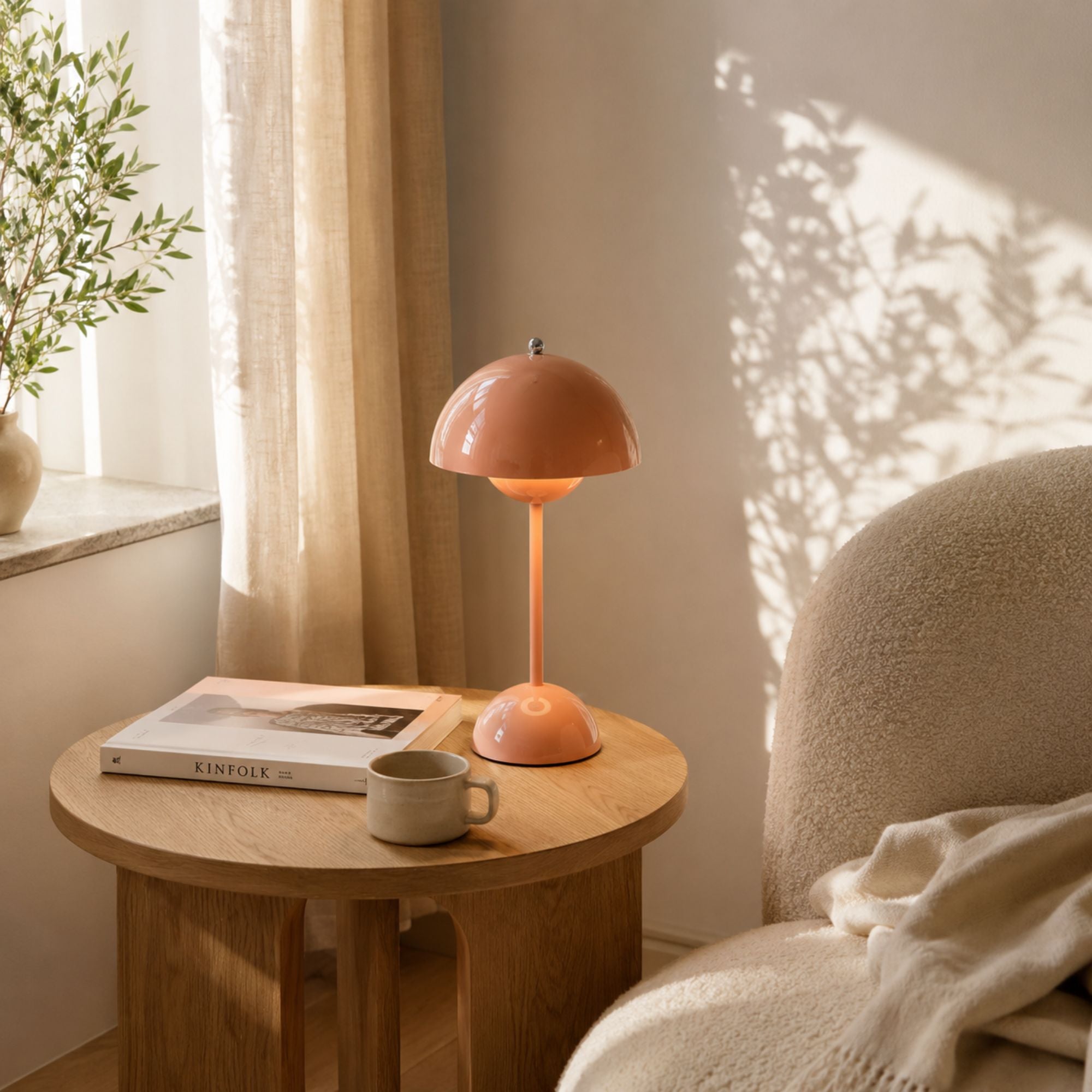 Rosa uppladdningsbar bordslampa med blank finish på runt ekbord i skandinavisk inredning. Omgiven av Kinfolk-magasin, keramikkopp och boucléfåtölj i mjukt dagsljus från fönster med linnégardiner.
