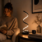 Modern svart spiralformad LED-bordslampa med varm stämningsfull belysning bredvid en kvinna som läser i en mysig och stilren kvällsmiljö med skandinavisk heminredning.