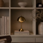 Portabel bordslampa i guld placerad i bokhylla med stilren skandinavisk inredning.