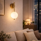 Modern vägglampa i guld med vit glaskupa ovanför soffa i mysigt vardagsrum på kvällen med varm och behaglig belysning